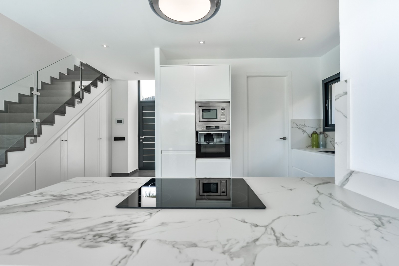 Moderne Küche mit marmorierter Arbeitsplatte und Induktionskochfeld, angrenzend an eine Treppe mit Glasgeländer und maßgefertigten Einbauschränken.