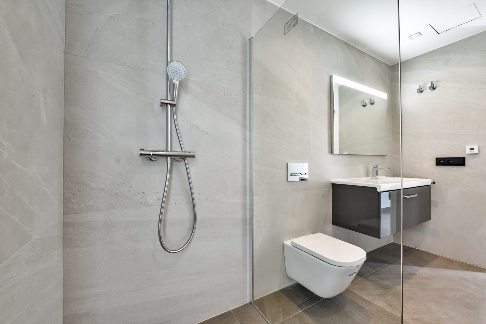Modernes Badezimmer mit begehbarer Dusche, Glastrennwand, wandhängendem WC und schwebendem Waschtisch. Großformatige graue Steinfliesen und hinterleuchteter Spiegel.