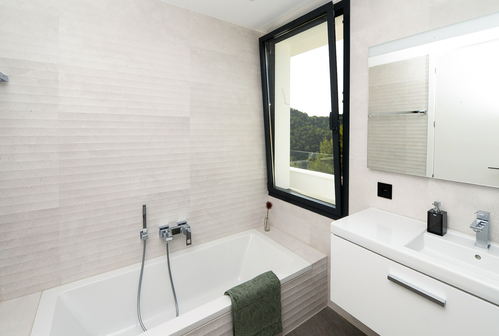 Modernes Badezimmer mit Einbaubadewanne, strukturierten Wandfliesen und Fenster mit Ausblick auf die bewaldete Hügellandschaft.