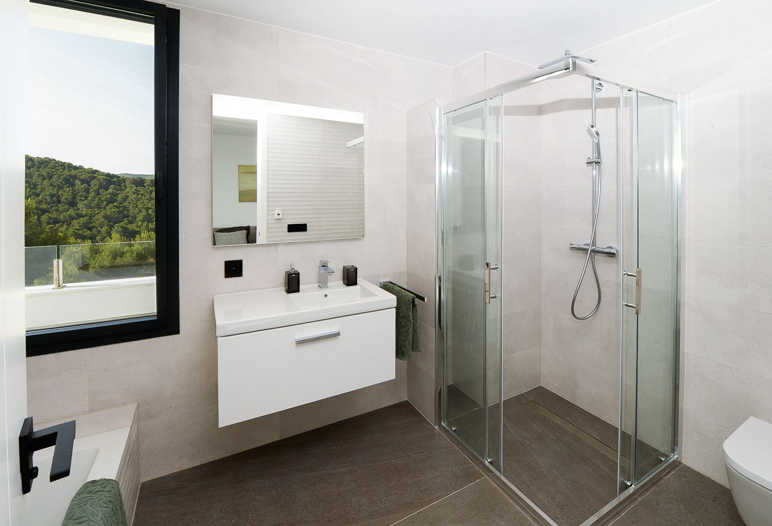 Modernes Badezimmer mit Glasduschkabine, weißem Hängewaschtisch und Fenster mit Blick auf die grünen Hügel. Neutrale Wandfliesen und dunkler Boden.