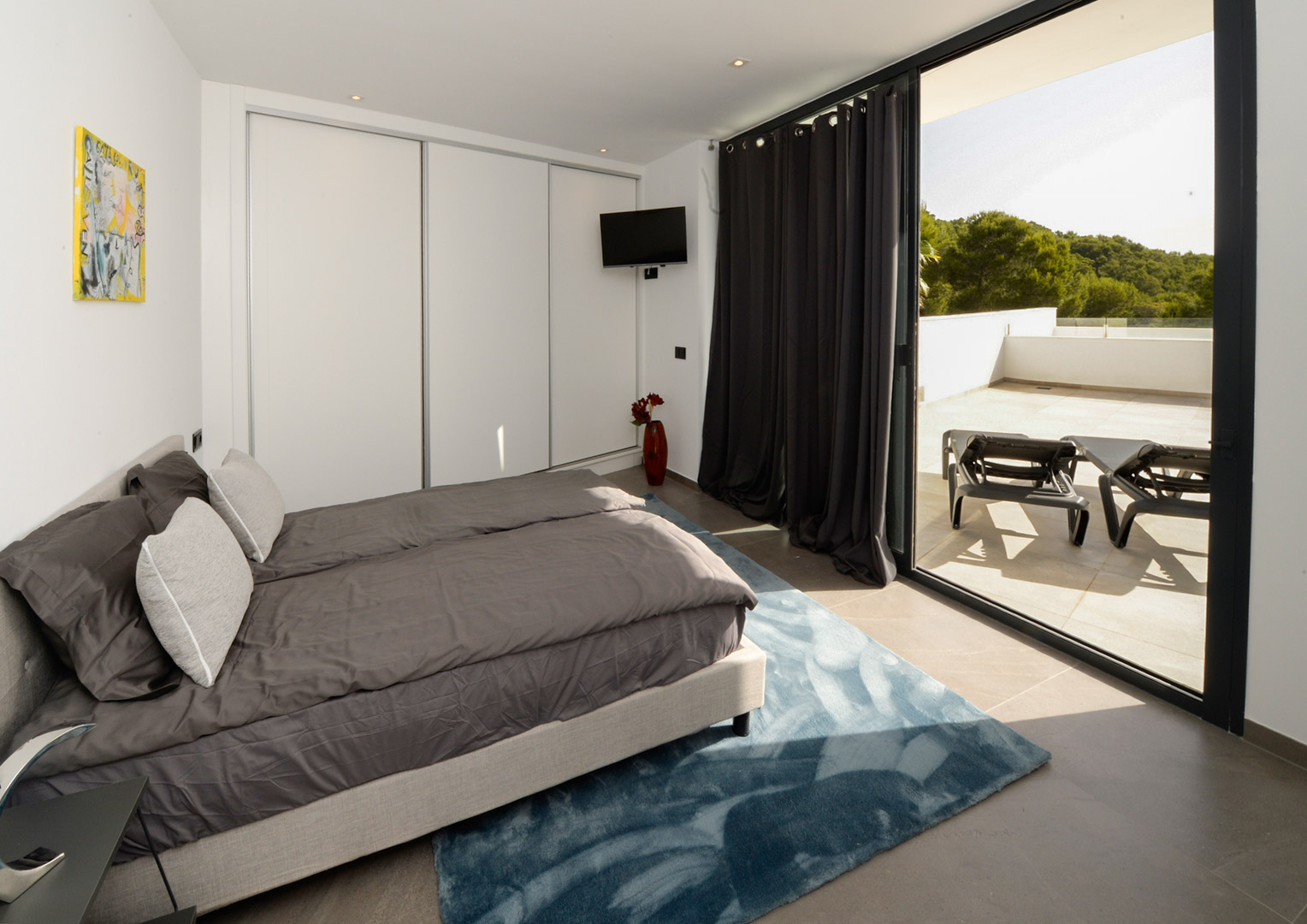 Modernes Schlafzimmer mit weißen Einbauschränken und direktem Zugang zu einer großen Terrasse mit Sonnenliegen und Blick in die grüne Umgebung.