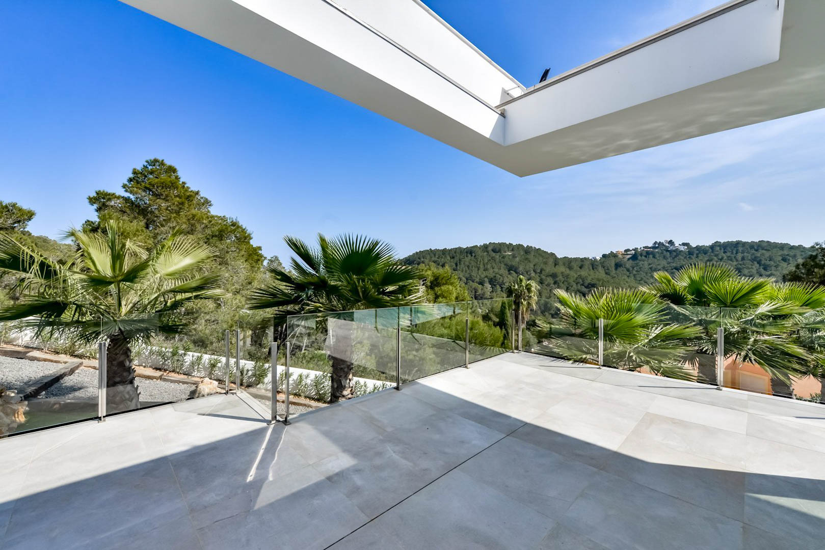 Obere Terrasse mit grauen Steinfliesen, Glasbalustraden und freiem Blick auf die bewaldeten Hügel und die umliegende mediterrane Vegetation.