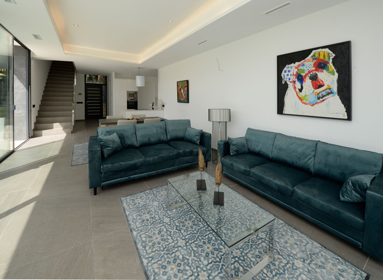 Offener Wohnbereich mit blaugrünen Samtsofas, Glastisch und grauen Bodenfliesen. Das Design umfasst indirekte Deckenbeleuchtung und einen nahtlosen Übergang zur Treppe.