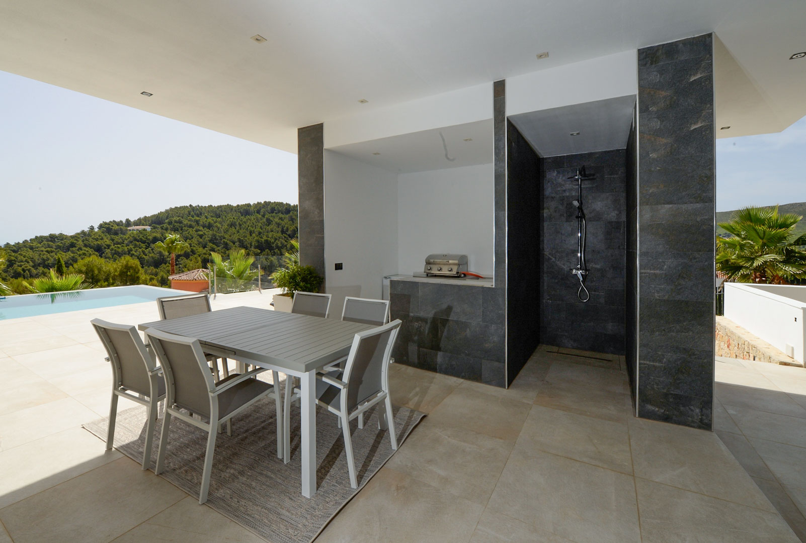 Überdachte Terrasse mit Essbereich, Außenküche mit Grill und einer mit dunklem Naturstein verkleideten Außendusche neben dem Poolbereich.