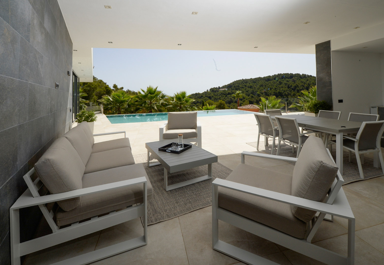 Überdachte Terrasse mit modernem Loungebereich und Essplatz im Freien, mit Blick auf den Infinity-Pool und die umliegende Hügellandschaft.