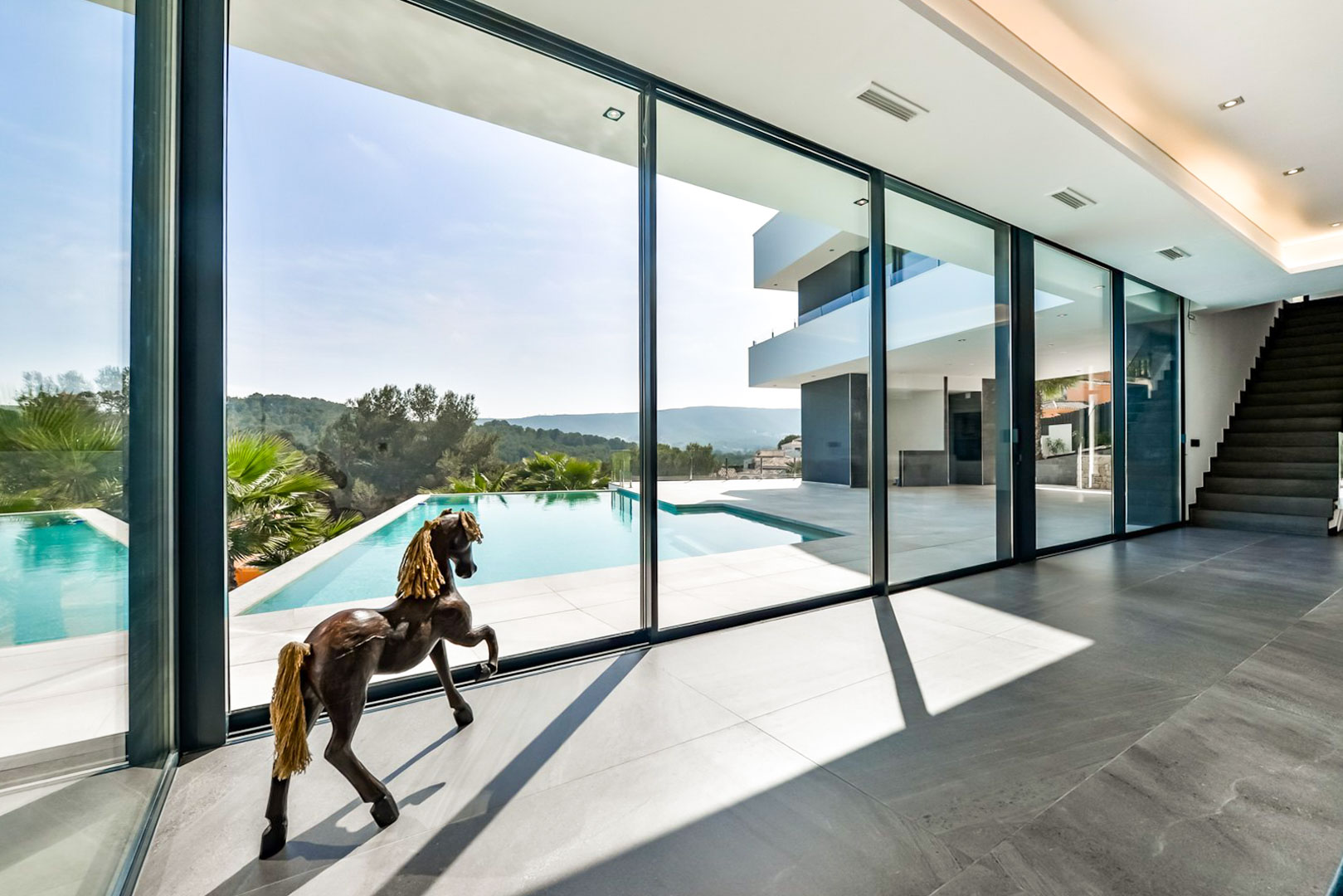 Wohnbereich mit bodentiefen Glasfronten, die den Blick auf den Infinity-Pool freigeben. Graue Feinsteinzeugböden und eine moderne Treppe prägen das offene Design.