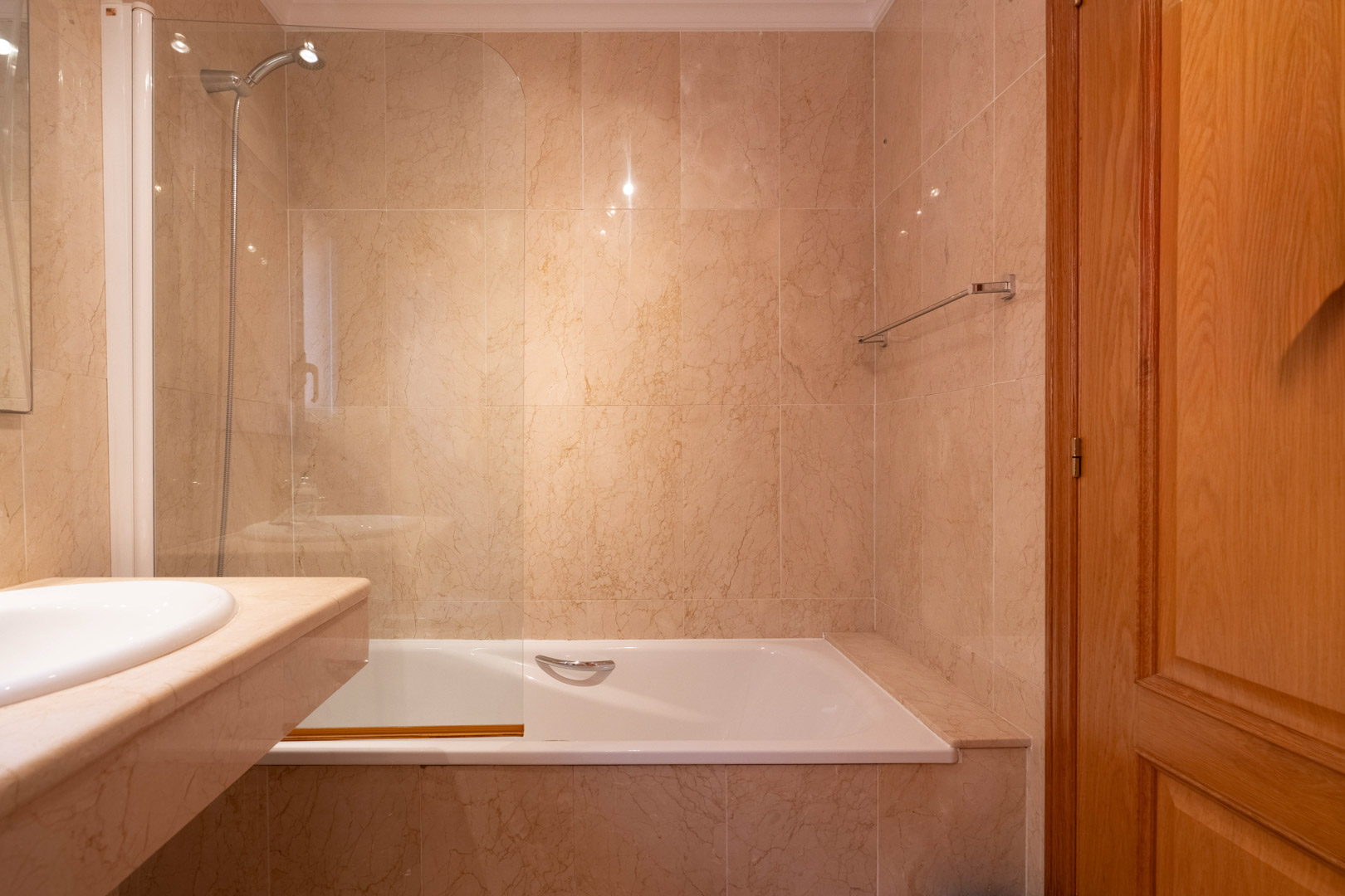 Badezimmer mit Badewanne, Glasduschwand und cremefarbenen Marmorwänden. Ausgestattet mit einem Waschtisch aus Naturstein und einer hochwertigen Holztür.