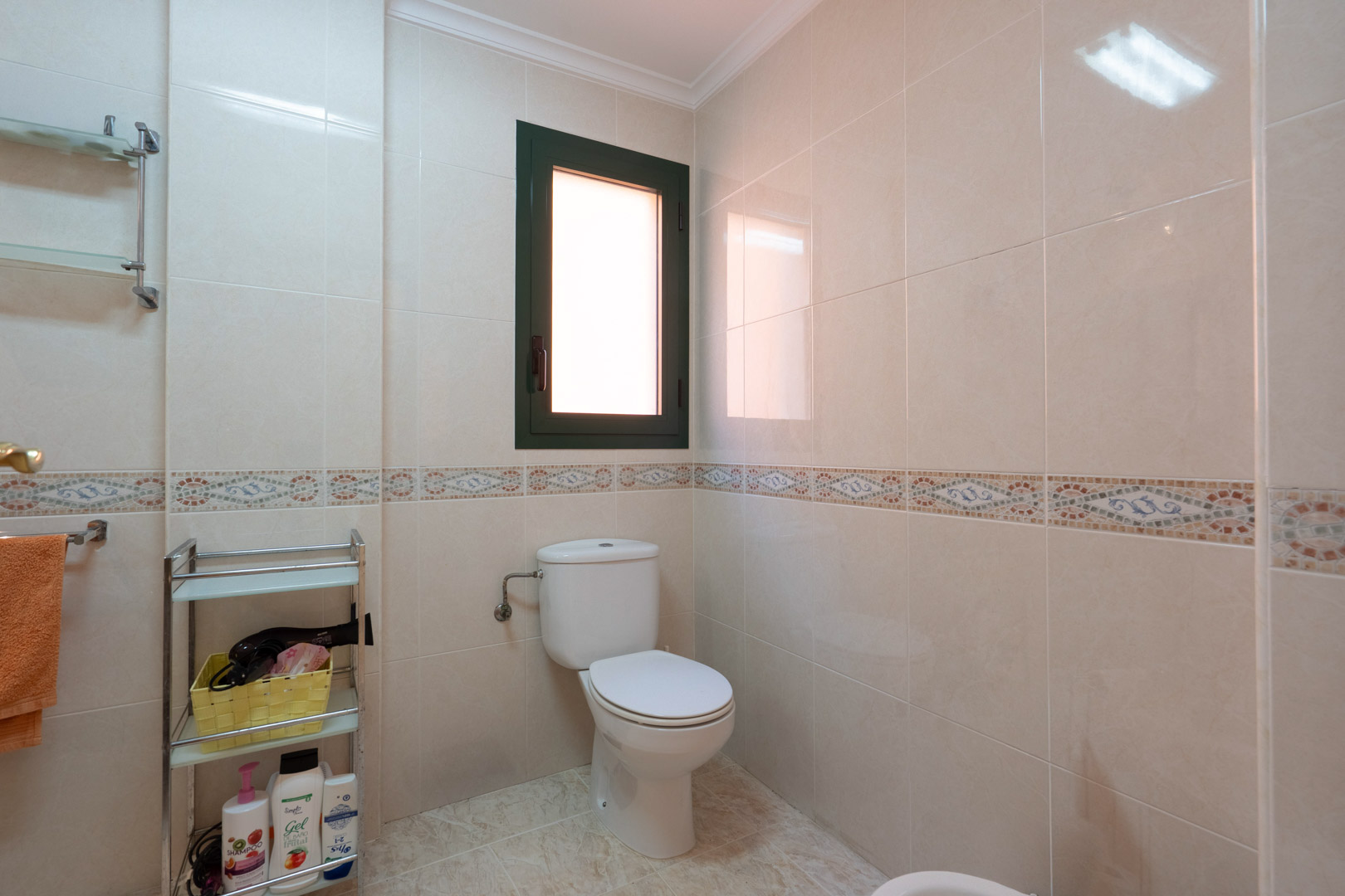 Badezimmer mit cremefarbenen Wandfliesen, dekorativer Mosaikbordüre, weißem WC und einem Fenster für natürliches Licht und Belüftung.