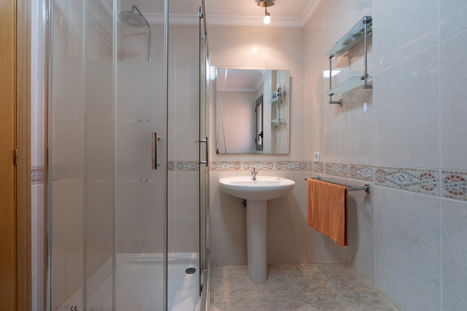 Funktionelles Badezimmer mit verglaster Dusche, Säulenwaschbecken und hell gefliesten Wänden mit einer umlaufenden Mosaikbordüre.