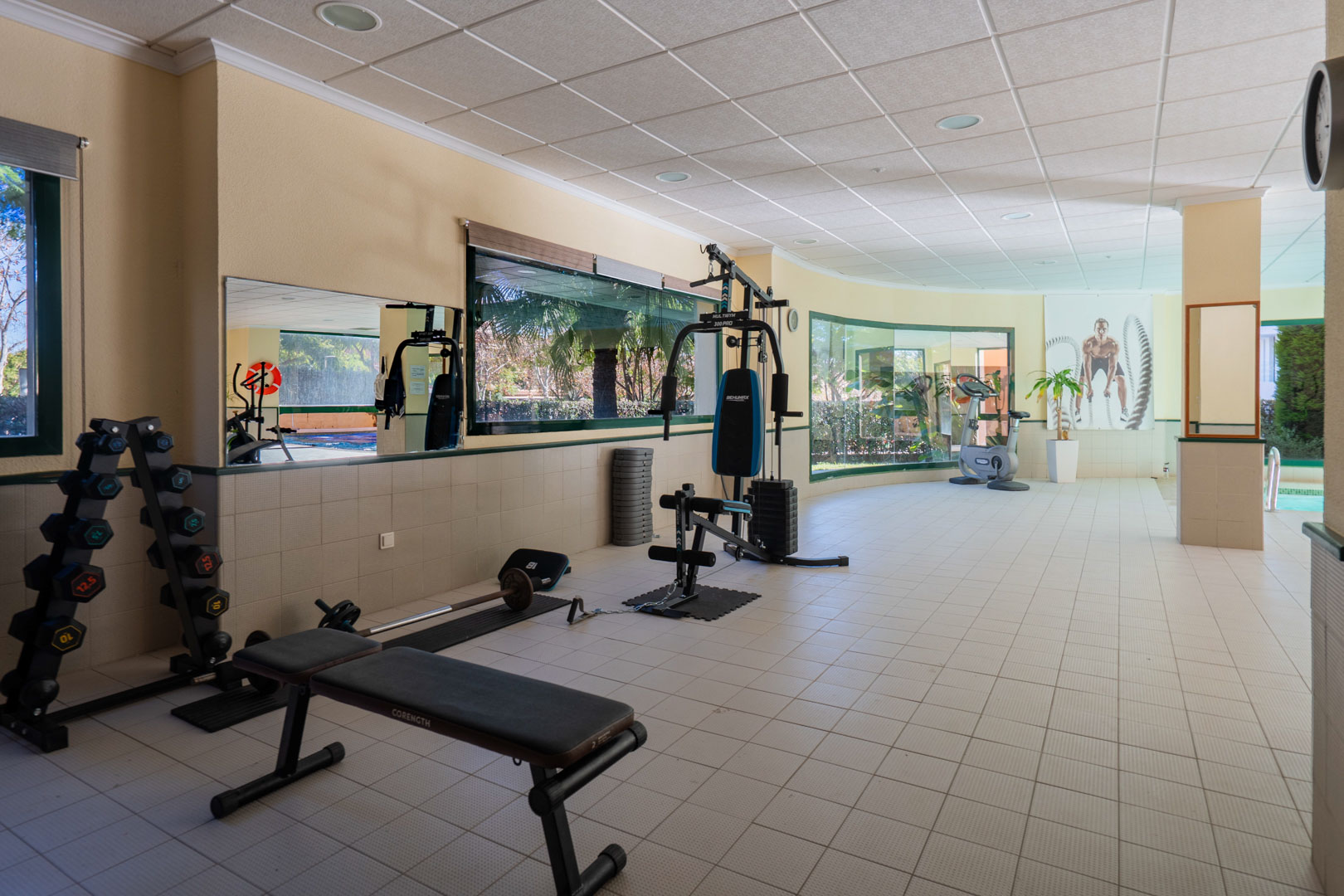 Hauseigenes Fitnessstudio mit Kraftgeräten, Hanteln und Kardiogeräten, hell gestaltet durch große Fensterfronten mit Blick auf die gepflegte Anlage.