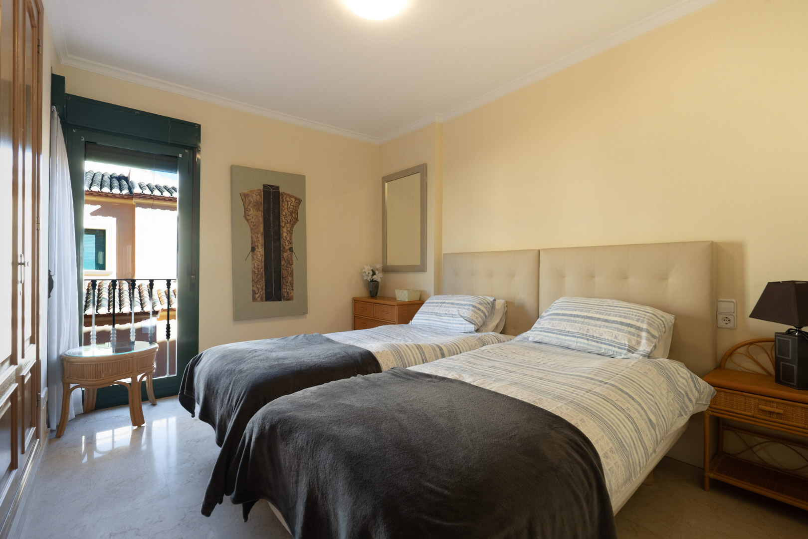 Helles Schlafzimmer mit zwei Einzelbetten, gepolstertem Kopfteil, Marmorboden und Einbauschränken. Direkter Zugang zu einem privaten Balkon.