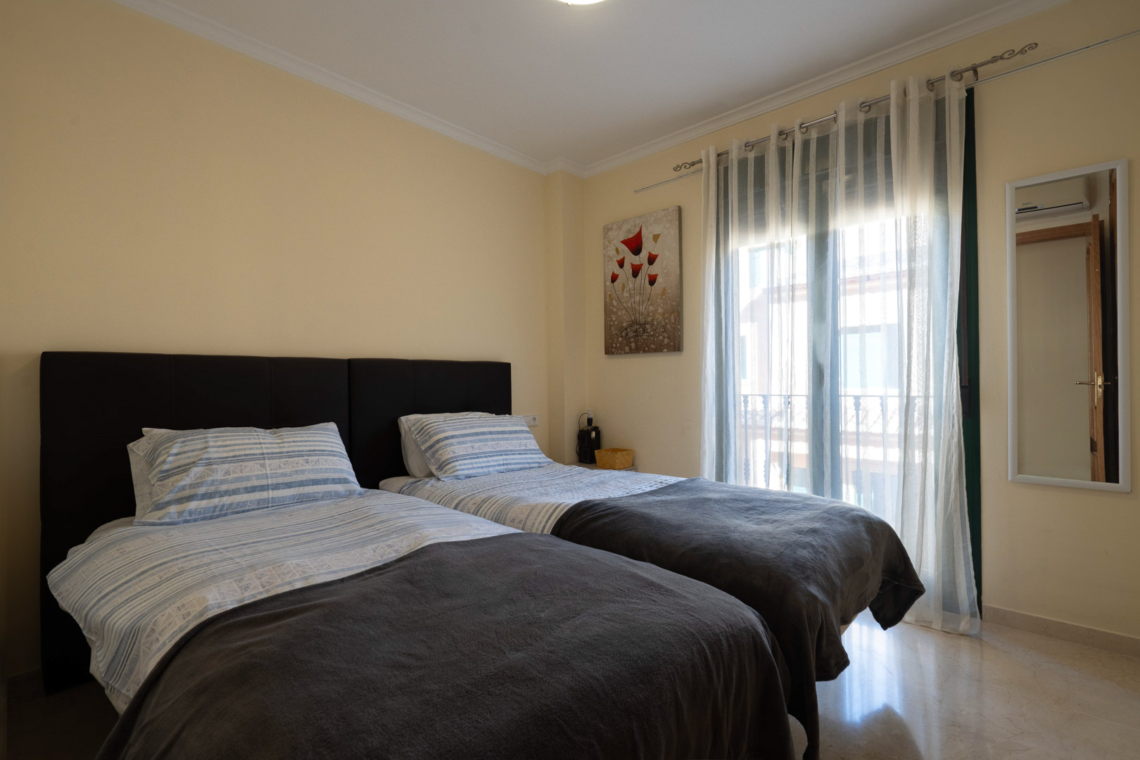 Helles Schlafzimmer mit zwei Einzelbetten, poliertem Marmorboden und Zugang zum Balkon durch bodentiefe Fenster.