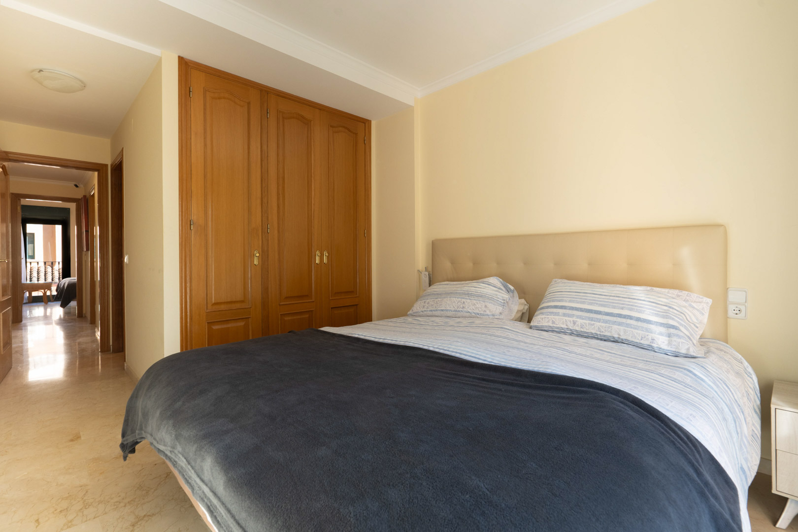 Schlafzimmer mit Einbauschränken aus Holz, Marmorboden und einem gepolsterten Kopfteil in neutralen Tönen, das an den hellen Flur grenzt.