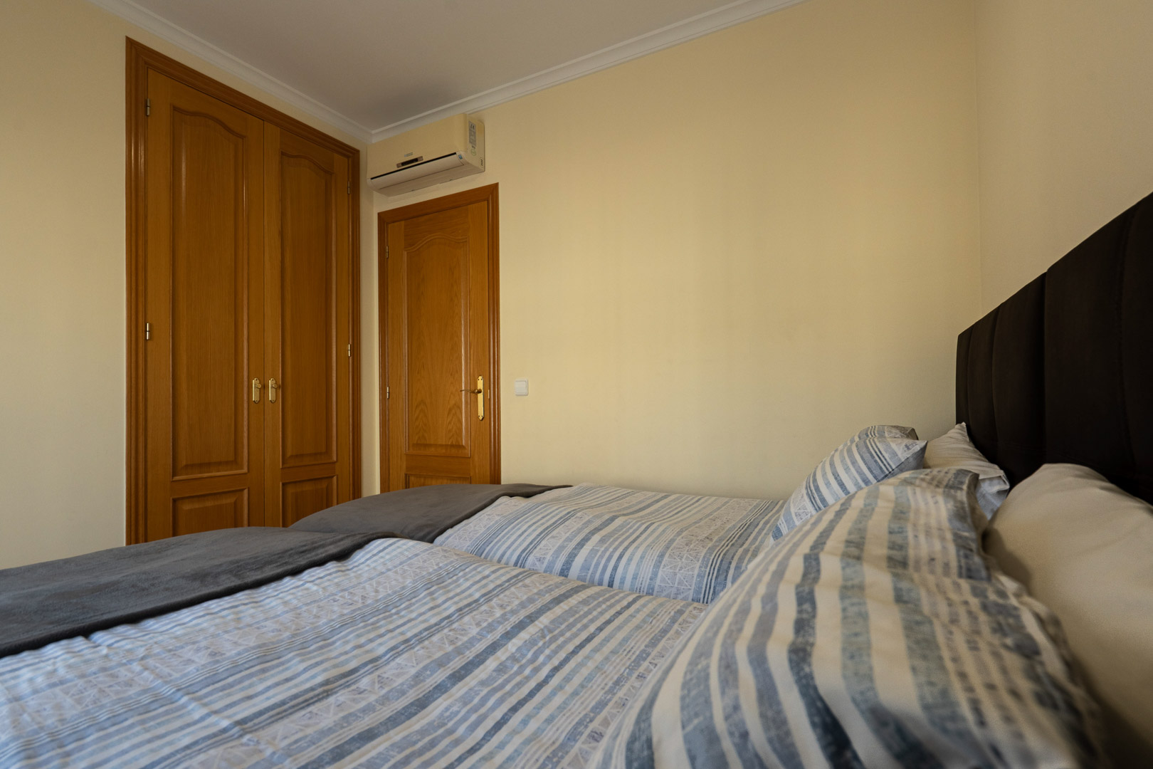 Schlafzimmer mit zwei Einzelbetten, Einbauschränken aus Holz und Split-Klimaanlage. Cremefarbene Wände und klassische Deckenleisten runden den Raum ab.