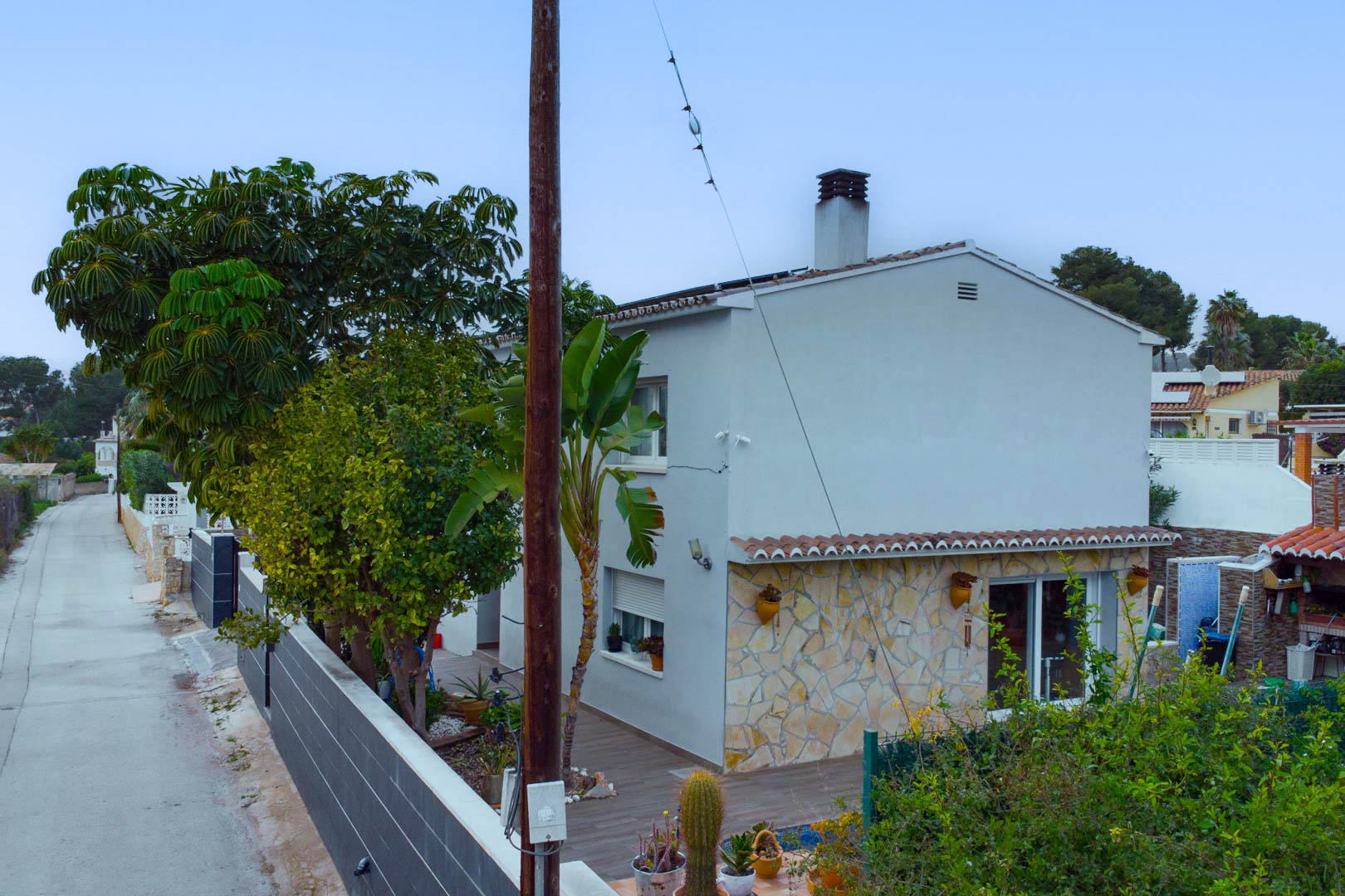Außenansicht der Villa mit weißer Fassade und Natursteinverkleidung, ergänzt durch mediterrane Bepflanzung und Solarmodule auf dem Dach.