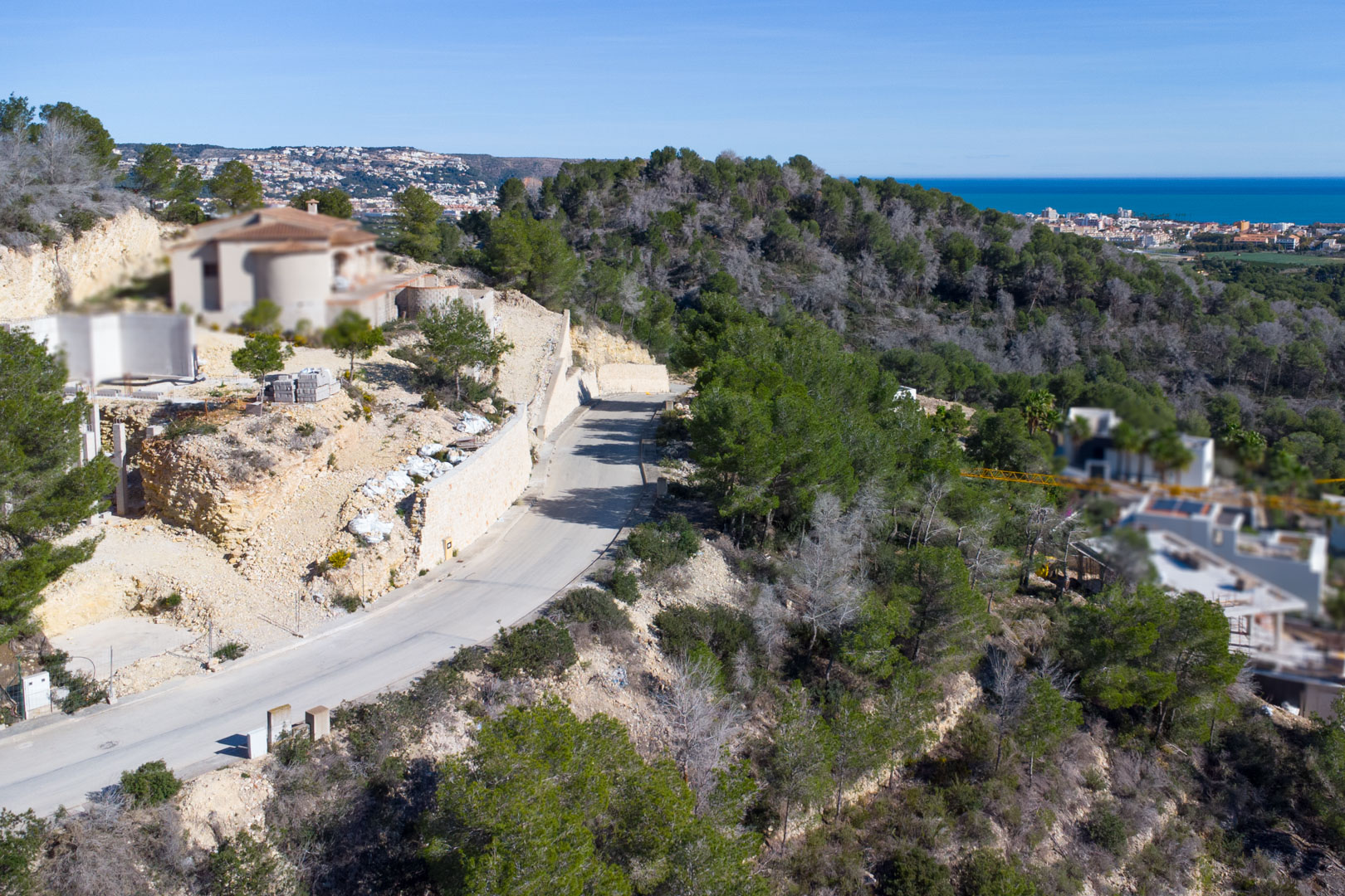 Luftaufnahme der asphaltierten Zufahrtsstraße in Tosalet 5 mit Stützmauern aus Naturstein, Pinienbewuchs und Blick auf das Mittelmeer und die Küste von Jávea.