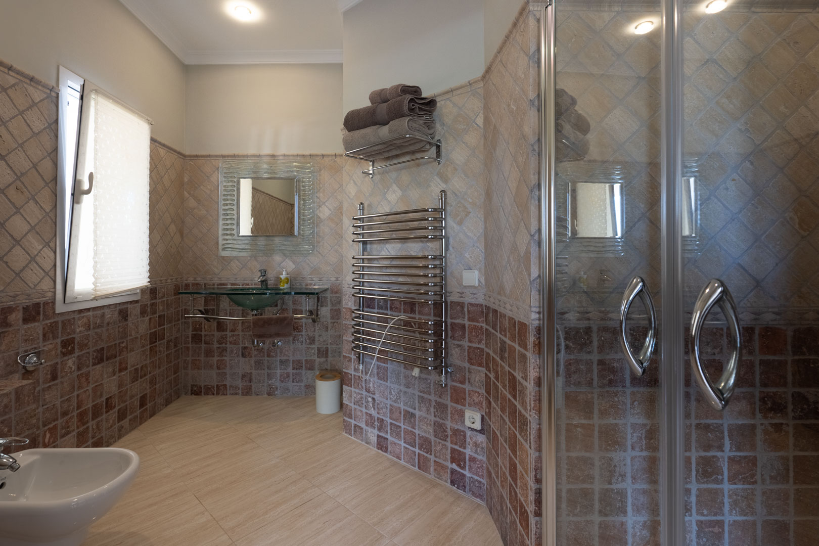 Badezimmer mit erdfarbenen Wandfliesen, einem gläsernen Waschbecken, verchromtem Handtuchheizkörper und einer Duschkabine aus Glas.