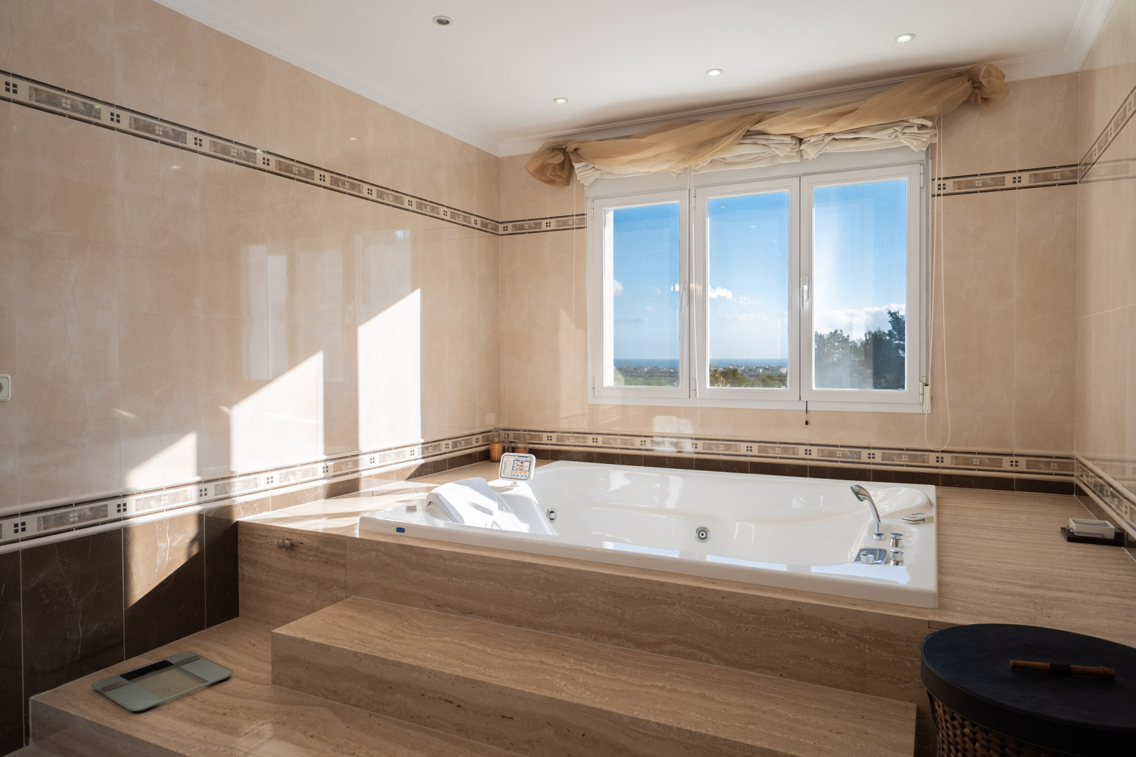 Badezimmer mit in Marmor eingefasster Hydromassage-Badewanne, großem Fenster mit Meerblick und eleganten beigen Fliesen mit dekorativen Bordüren.