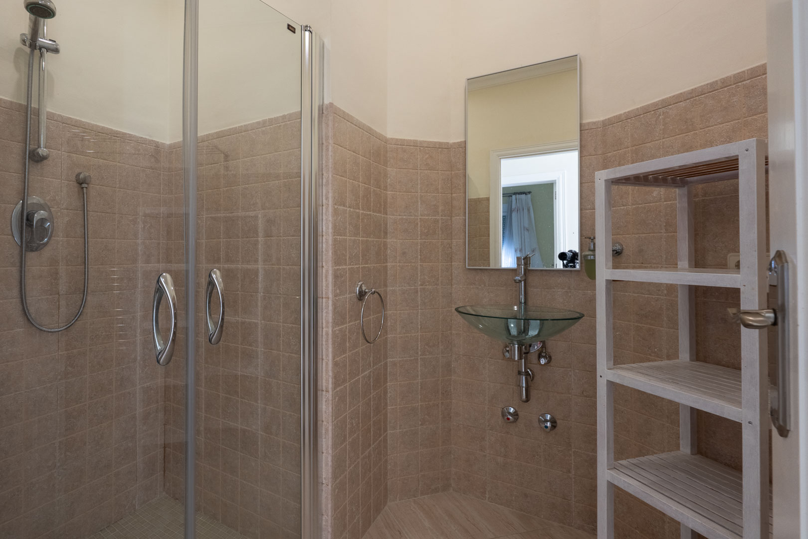 Badezimmer mit verglaster Dusche, einem hängenden Glaswaschbecken, beigefarbenen Wandfliesen und einem weißen Holzregal für zusätzlichen Stauraum.