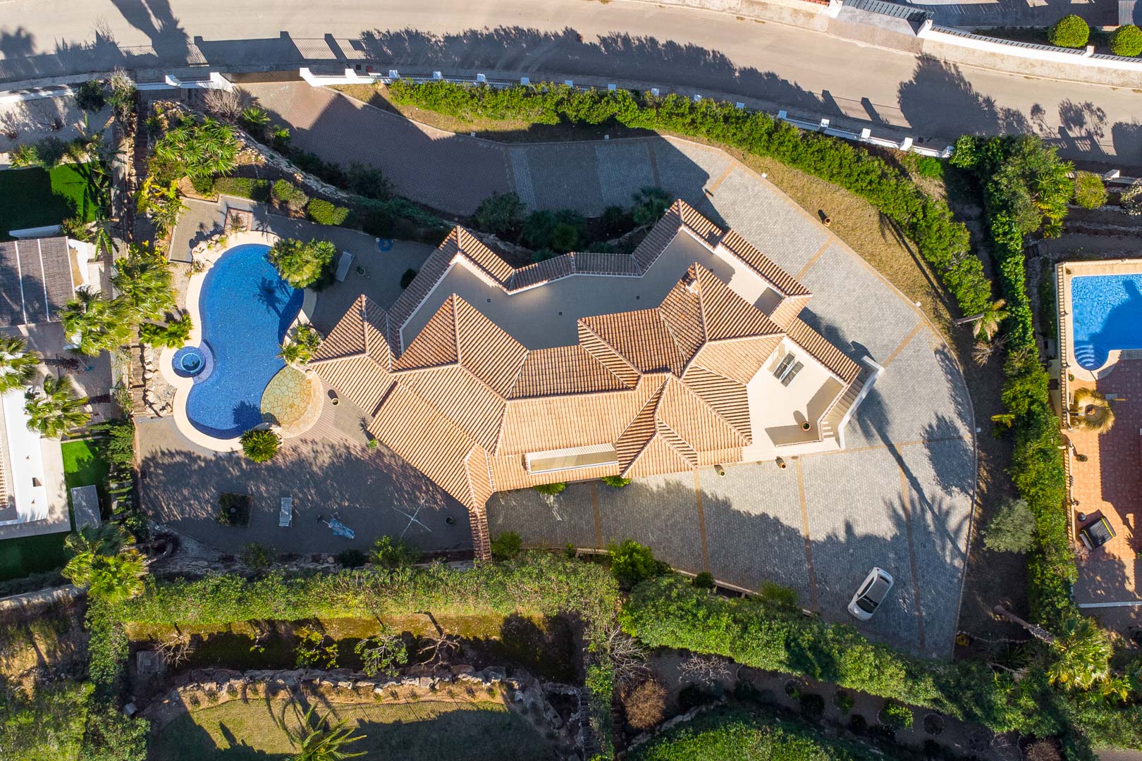 Draufsicht aus der Vogelperspektive auf das Grundstück mit seinem mehrteiligen Ziegeldach, dem nierenförmigen Pool mit Jacuzzi und der großzügig gepflasterten Einfahrt.