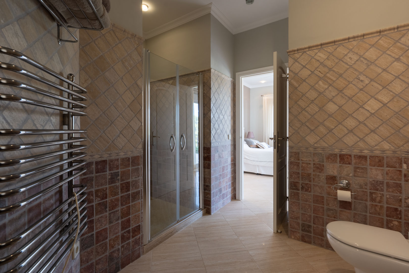 En-Suite-Badezimmer mit Glasdusche, verchromtem Handtuchwärmer und zweifarbigem Fliesenspiegel, mit Blick in das angrenzende Schlafzimmer.