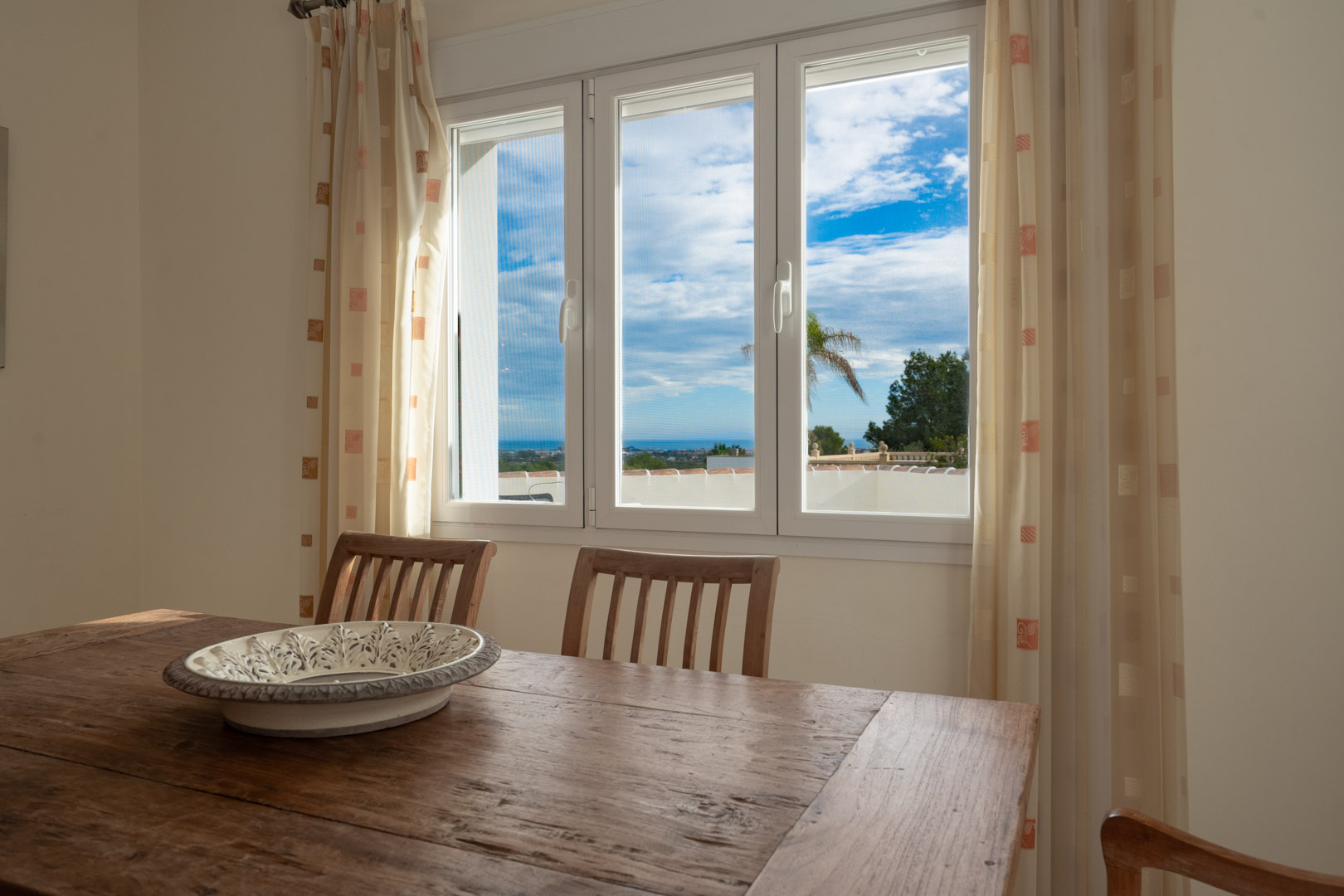 Essbereich mit rustikalem Holztisch und Blick auf das Mittelmeer durch ein weißes dreiflügeliges Fenster, das für helles, natürliches Licht sorgt.