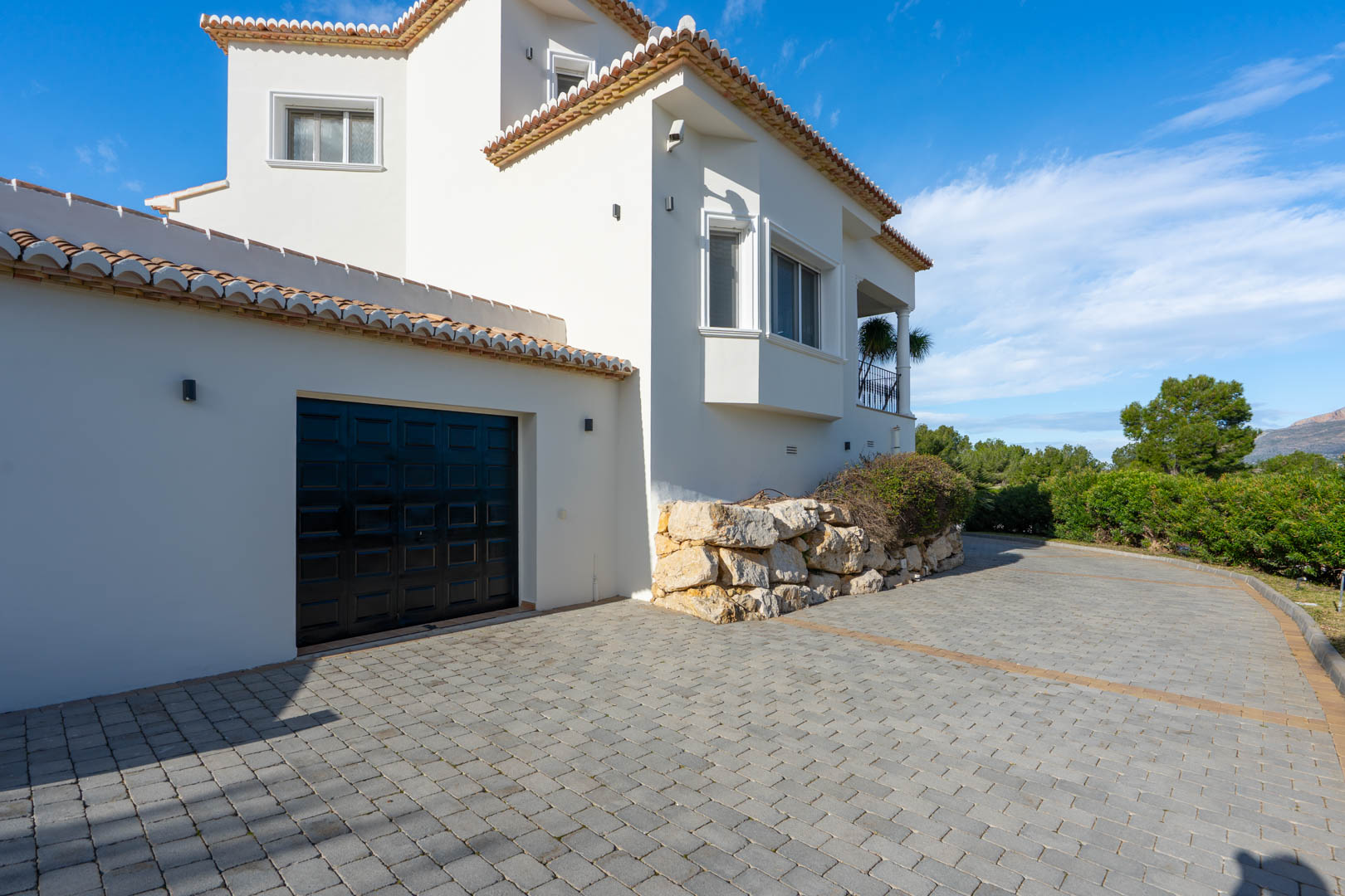Gepflasterte Einfahrt mit Zugang zur Garage, ergänzt durch eine weiße mediterrane Fassade und markante Stützmauern aus Naturstein unter blauem Himmel.