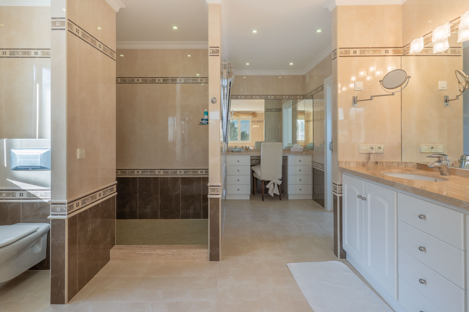 Großzügiges Badezimmer mit zweifarbigen Fliesen, begehbarer Dusche, Frisiertisch und Waschtisch mit Marmorplatte sowie weißen Unterschränken.