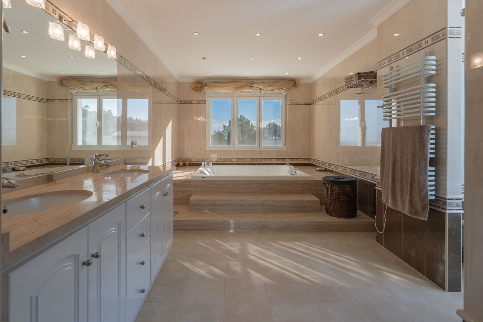 Hauptbadezimmer mit Doppelwaschtisch aus Marmor, erhöhter Whirlpool-Badewanne auf Steinstufen und hochwertigen Wandfliesen mit Zierbordüren.