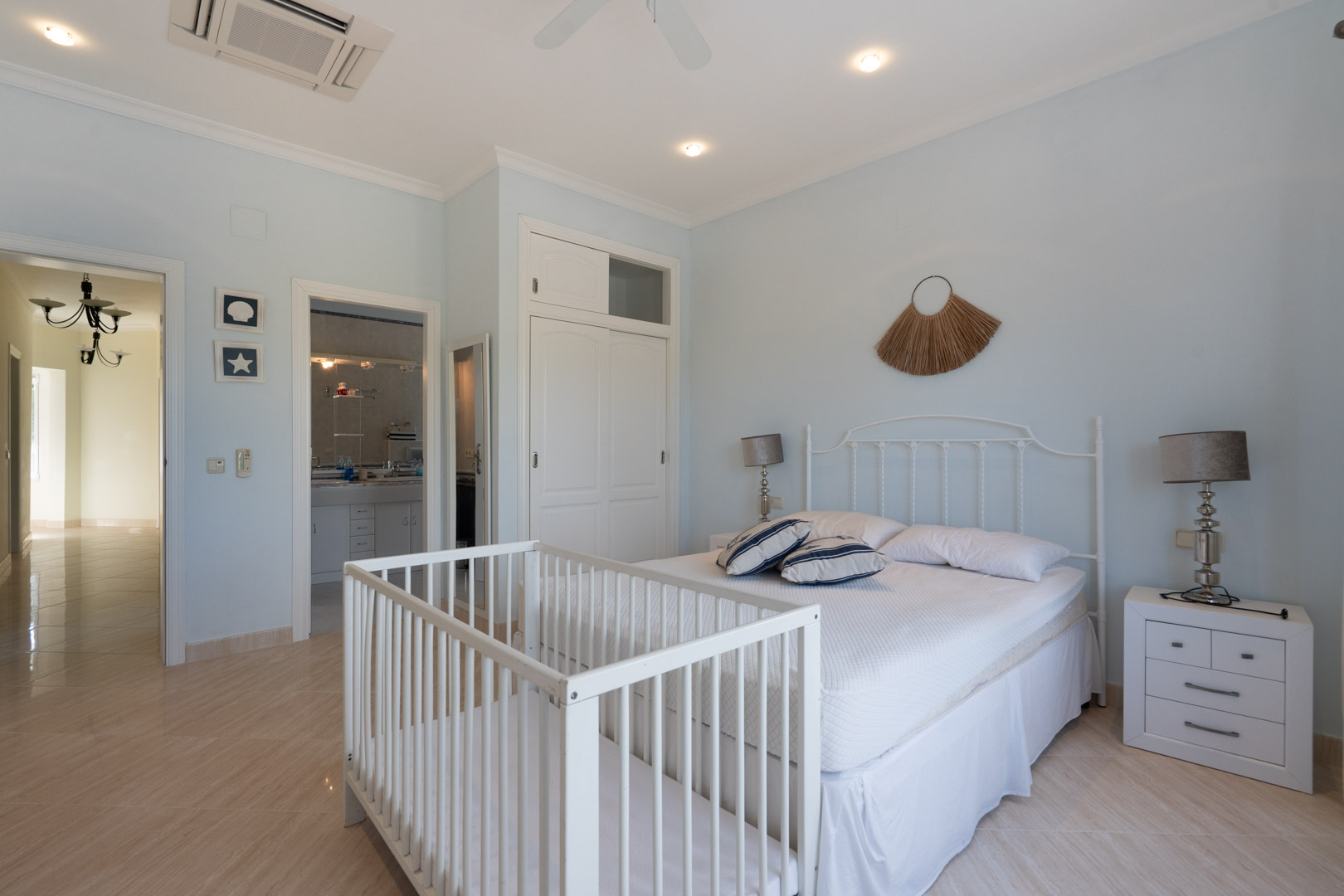 Helles Schlafzimmer mit blassblauen Wänden, weißem Schmiedeeisen-Kopfteil, Kinderbett, Einbauschränken und direktem Zugang zum En-Suite-Badezimmer.