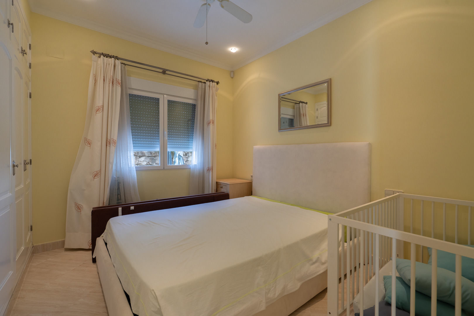 Helles Schlafzimmer mit cremefarbenen Wänden, weißen Einbauschränken, Doppelbett mit gepolstertem Kopfteil und Kinderbett. Ausgestattet mit Deckenventilator und Einbaustrahlern.