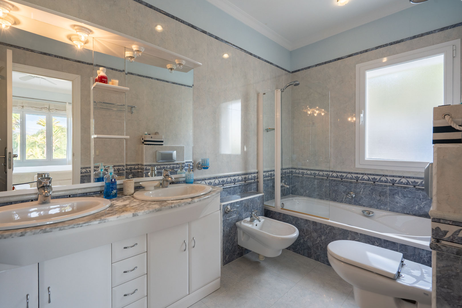 Komplettes Badezimmer mit Doppelwaschtisch auf Marmorplatte, Badewanne mit Glastrennwand und klassischen blau-grauen Fliesen mit dekorativen Bordüren.