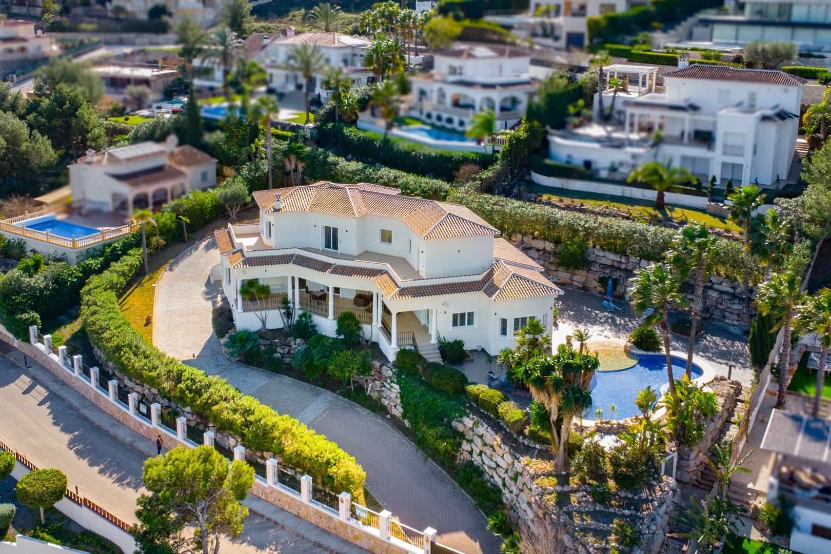 Luftaufnahme der Villa mit weißer Fassade und Terrakotta-Dach, großzügiger gepflasterter Einfahrt sowie einem Poolbereich inmitten mediterraner Bepflanzung.