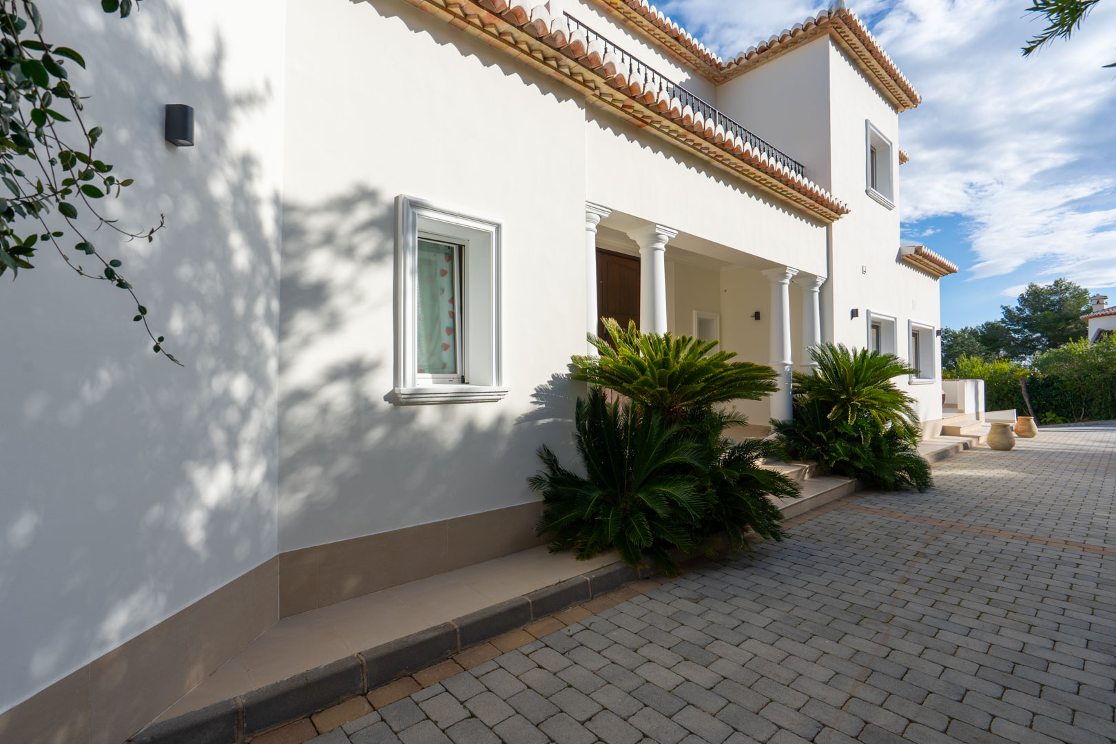 Mediterrane Fassade mit weißen Säulen, gepflasterter Einfahrt und architektonischen Details wie Terrakotta-Dachziegeln und gepflegter Bepflanzung.