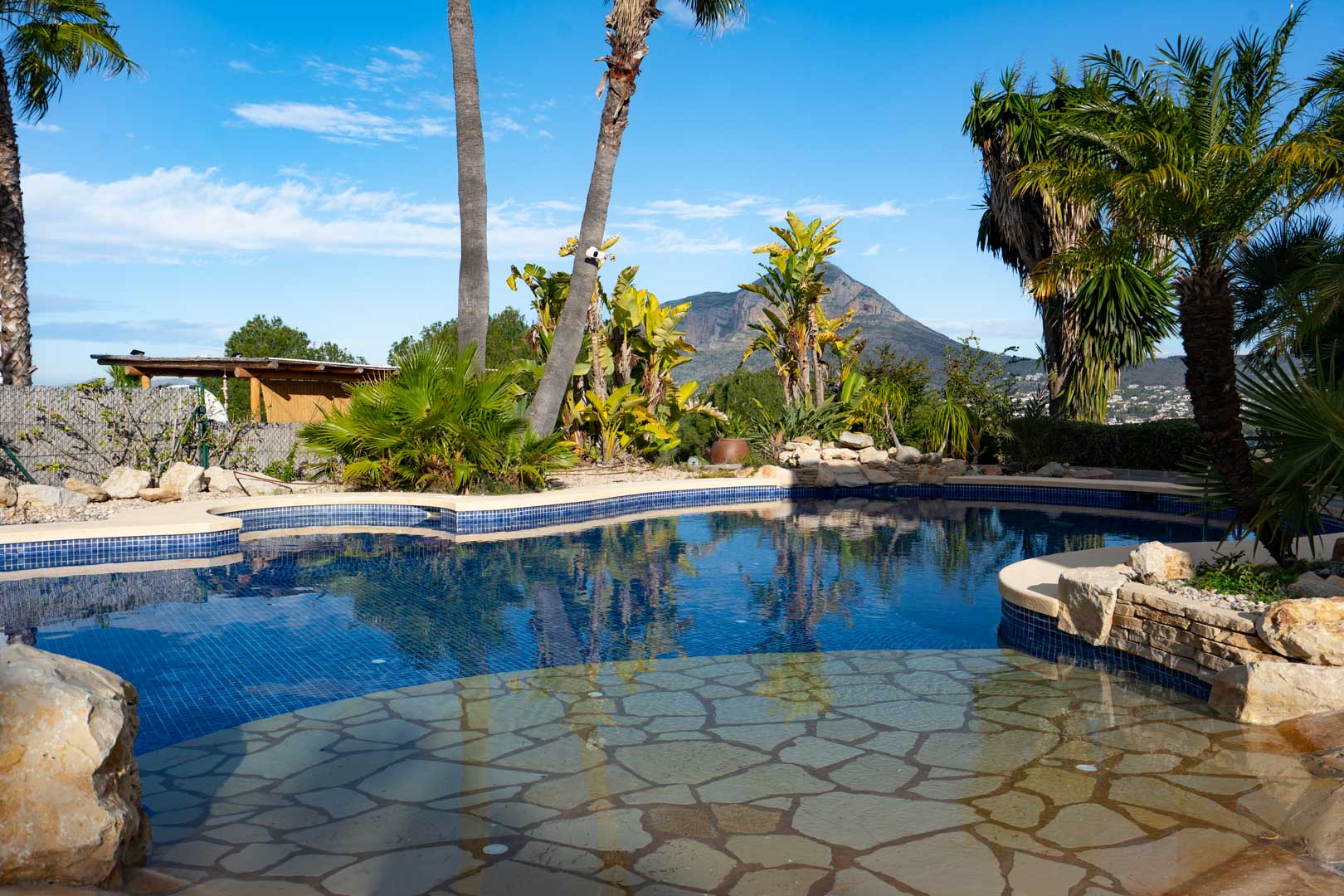 Organisch geformter Pool mit blauen Mosaikfliesen und flachem Naturstein-Einstieg, eingebettet in einen tropischen Garten mit Blick auf den Montgó.