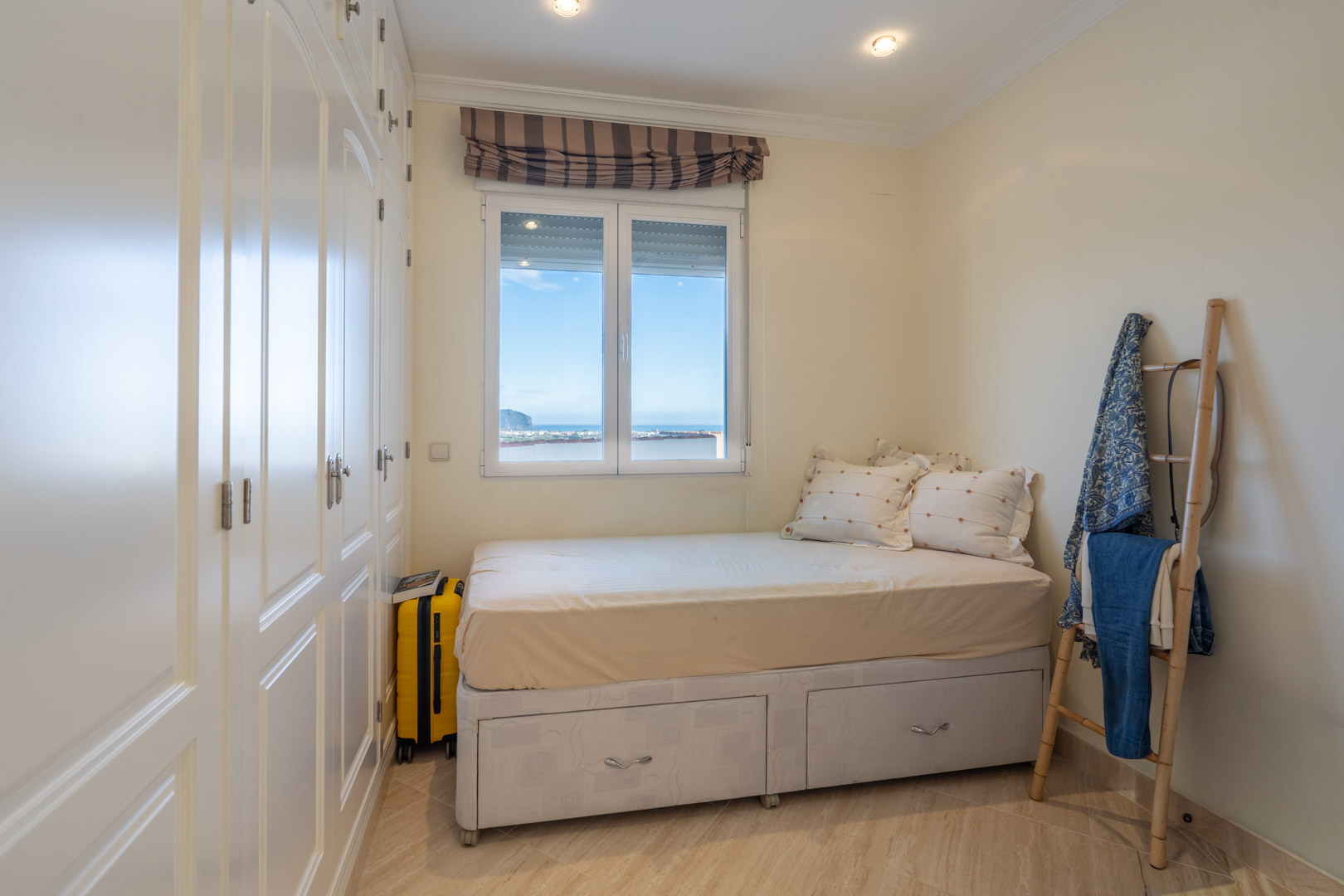 Schlafzimmer mit weißen Einbauschränken, Bett mit integrierten Schubladen und Fenster mit direktem Blick auf das Mittelmeer.