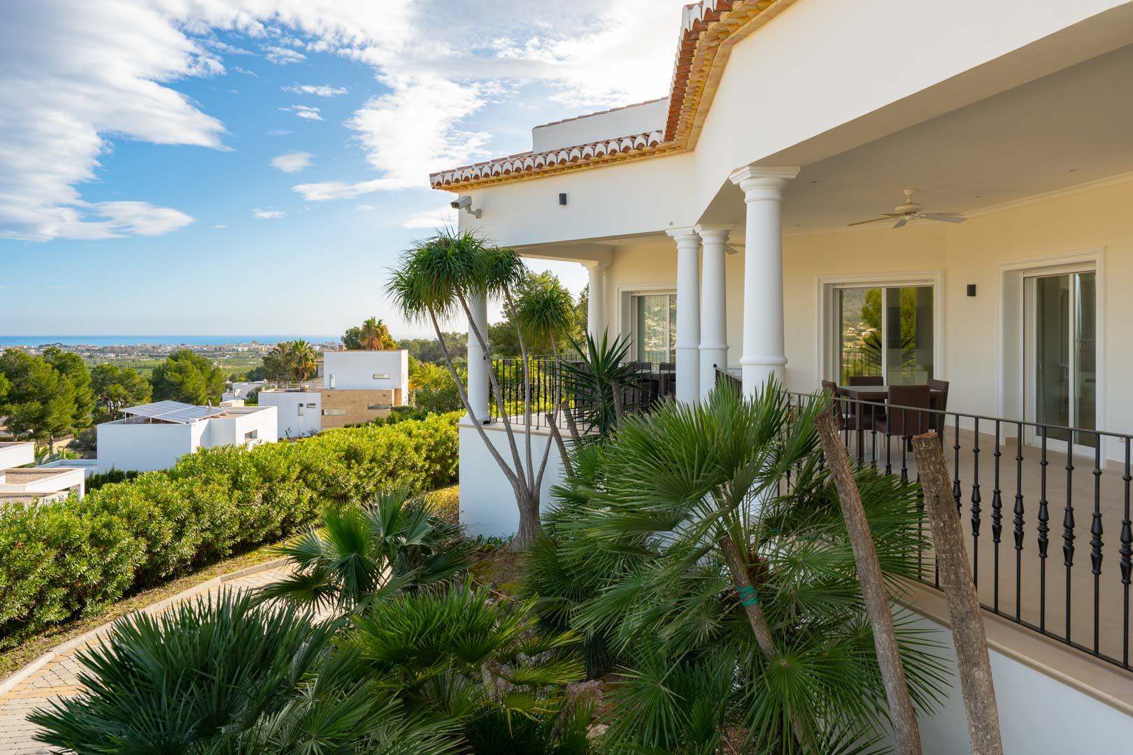 Überdachte Terrasse mit klassischen Säulen und Schmiedeeisengeländer, umgeben von tropischen Palmen und mit Panoramablick auf das Mittelmeer.