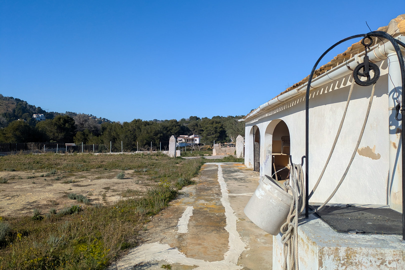 Außenansicht der Finca mit traditionellem Brunnen und Seilwinde, neben der weißen Bogenfassade und einem weitläufigen, flachen Naturgrundstück.