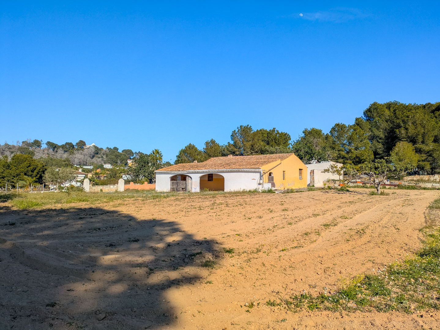 Außenansicht der traditionellen Finca mit Rundbogen-Veranda, Ziegeldach und einem weitläufigen, flachen Naturgrundstück inmitten von Pinien.