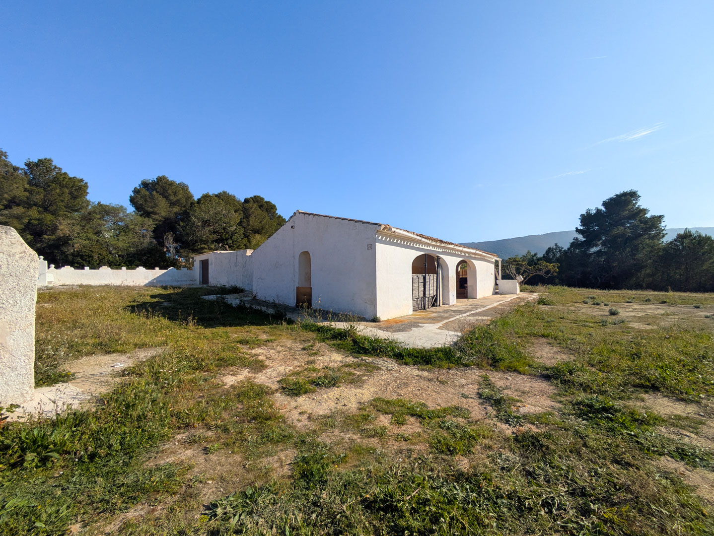 Außenansicht der traditionellen Finca mit weiß getünchten Wänden und Rundbögen auf einem weitläufigen, flachen Naturgrundstück mit Pinienbestand.