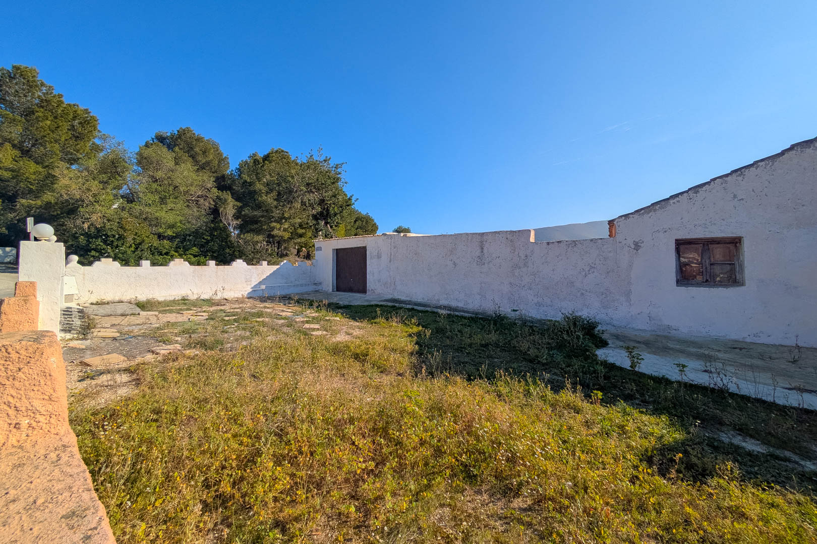 Außenansicht der weiß getünchten Fassade mit originalen Holzfenstern und ummauertem rustikalem Hof, umgeben von mediterranen Pinien und Vegetation.