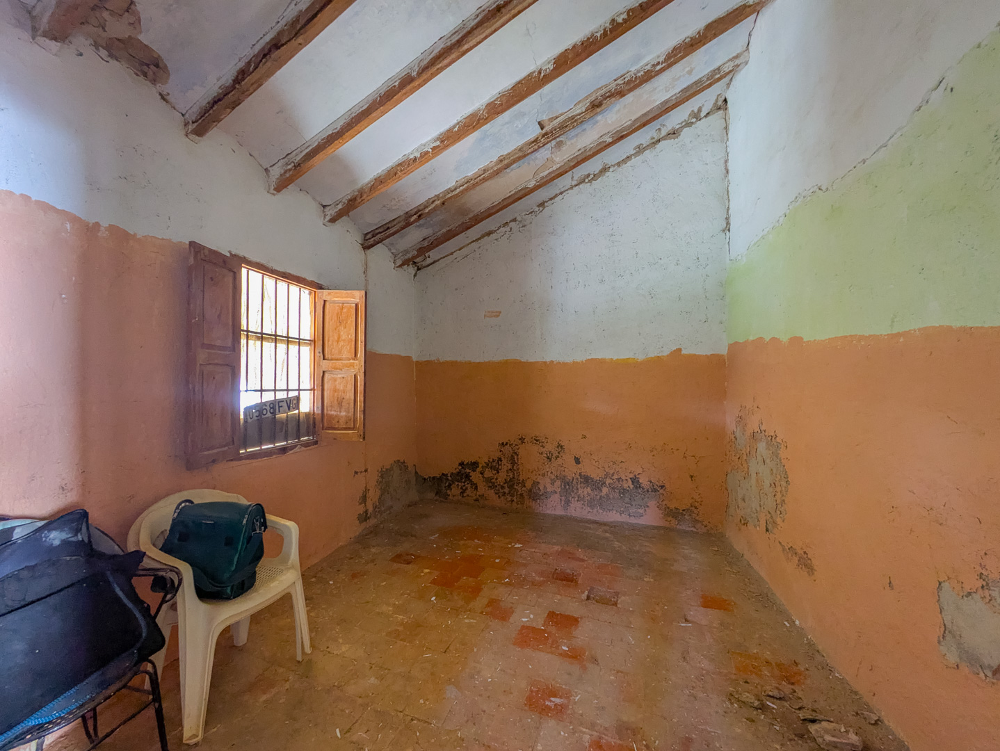 Innenraum mit schräger Decke und sichtbaren Holzbalken, traditionellem Terrakotta-Fliesenboden und einem kleinen Fenster mit originalen Holzläden.