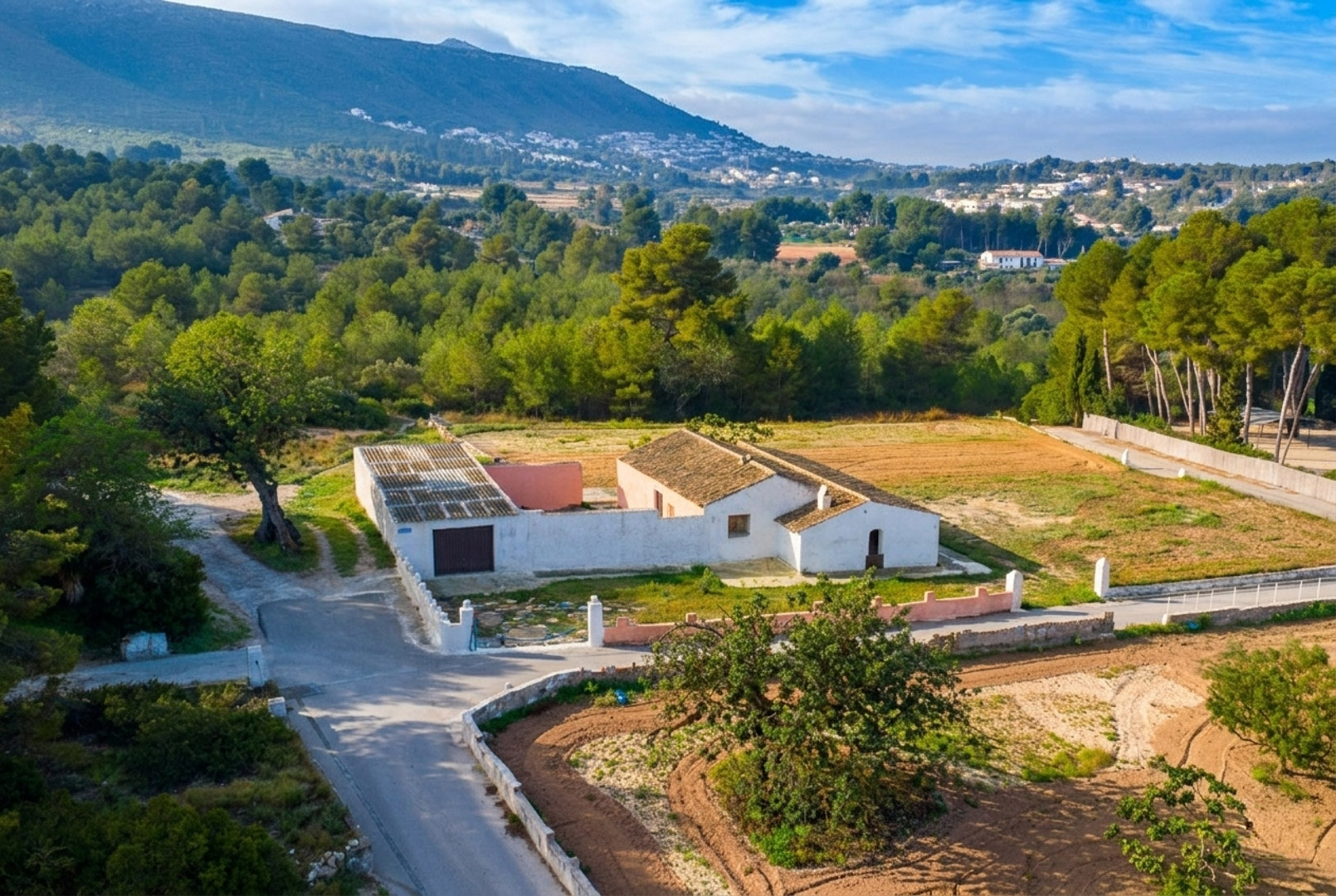 Luftaufnahme der traditionellen Finca mit weißen Mauern und Ziegeldächern auf einem flachen, rustikalen Grundstück, umgeben von Pinienwäldern und Bergen.