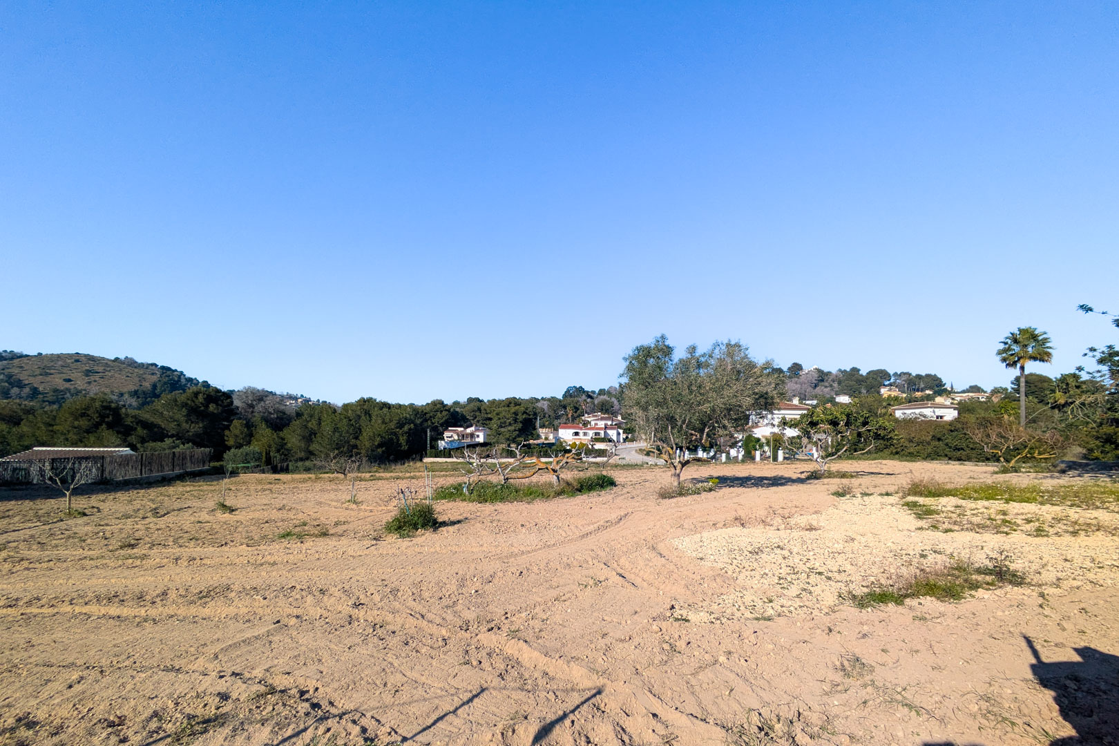 Panoramablick auf das weitläufige, flache rustikale Grundstück mit gepflügter Erde, mediterranen Bäumen und Blick auf die umliegenden Hügel unter blauem Himmel.