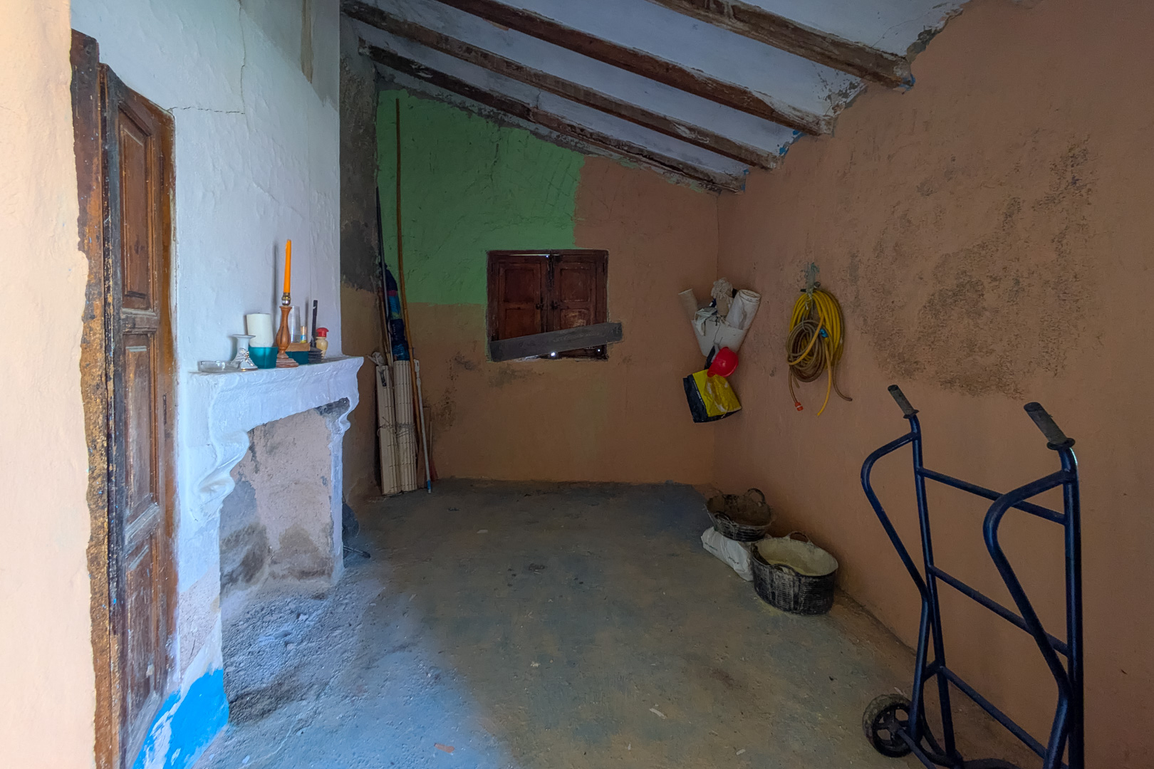 Rustikaler Innenraum mit traditionellem Kamin, schräger Holzbalkendecke und strukturierten Wänden, der das Renovierungspotenzial des Hauses zeigt.