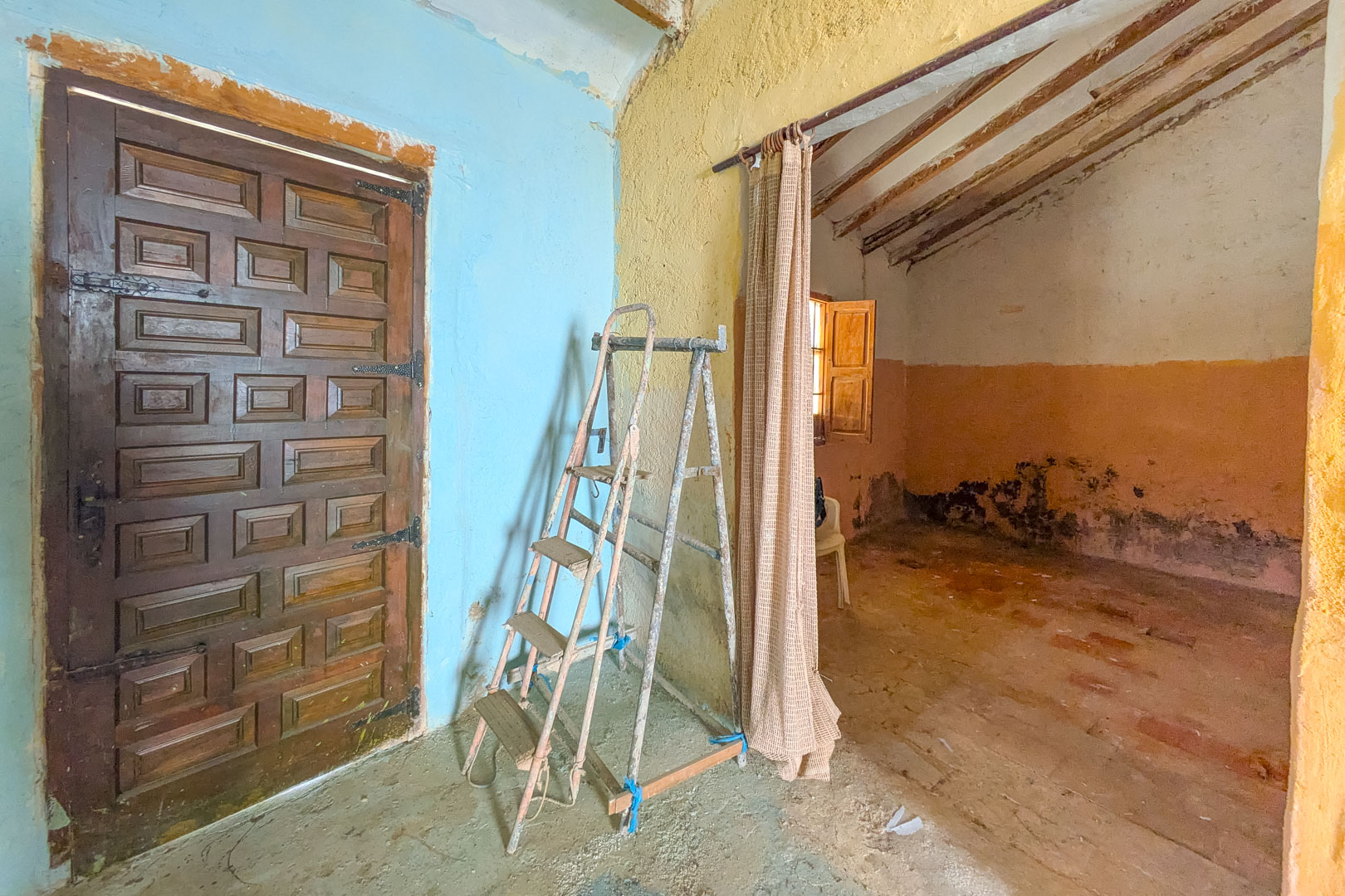 Rustikaler Innenraum mit traditioneller Holztür, farbigen Wänden und sichtbaren Deckenbalken, der das umfassende Renovierungspotenzial verdeutlicht.