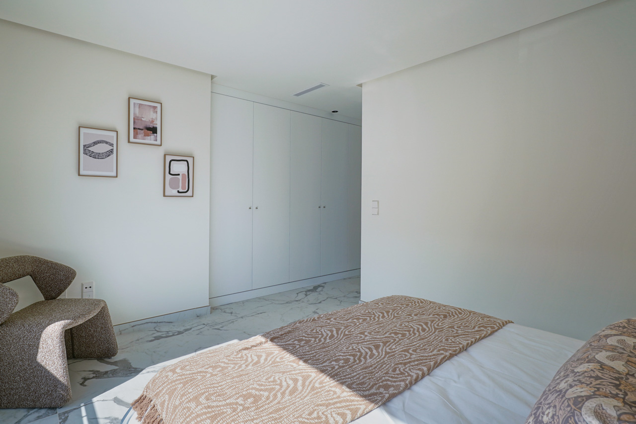 Helles Schlafzimmer mit raumhohen weißen Einbauschränken, Marmoreffekt-Boden und einer modernen Leseecke mit Designer-Sessel.