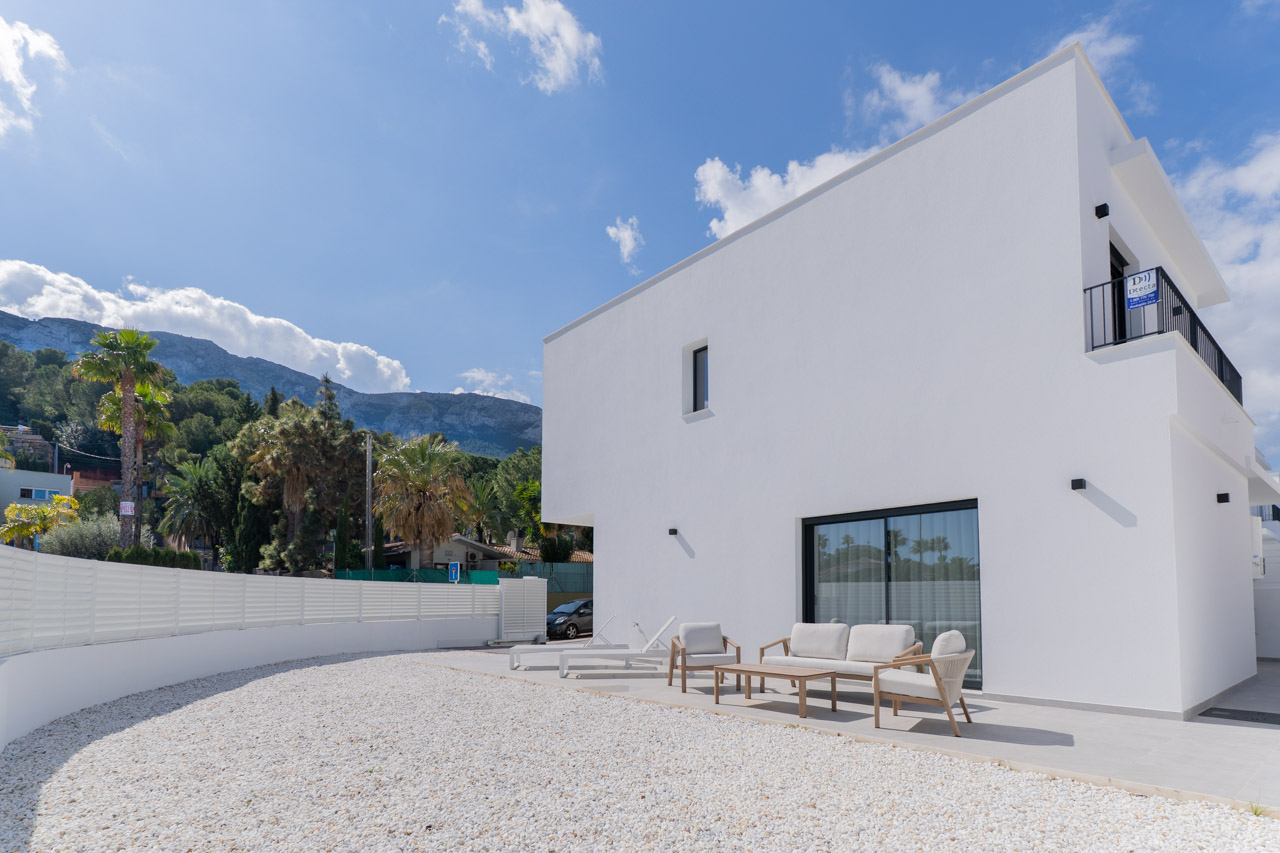 Minimalistische weiße Fassade mit gepflasterter Terrasse, Gartenmöbeln und weißem Kiesgarten, mit Blick auf das Montgó-Massiv im Hintergrund.