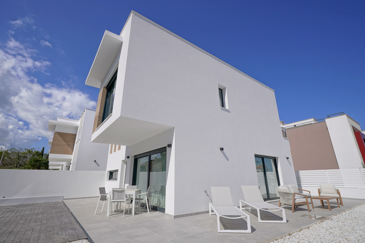 Moderne Außenansicht mit weißen geometrischen Formen, einer gefliesten Terrasse mit Gartenmöbeln und Sonnenliegen sowie einem privaten Parkplatz.