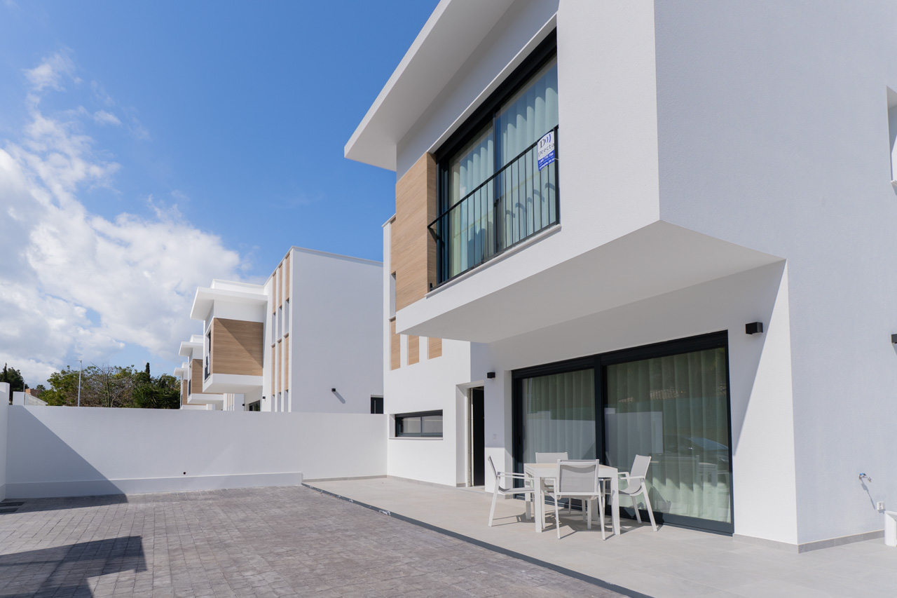 Moderne Außenansicht mit weißer Fassade, Holzoptik-Elementen, gepflasterter Terrasse und Essbereich im Freien vor großen Glasschiebetüren.